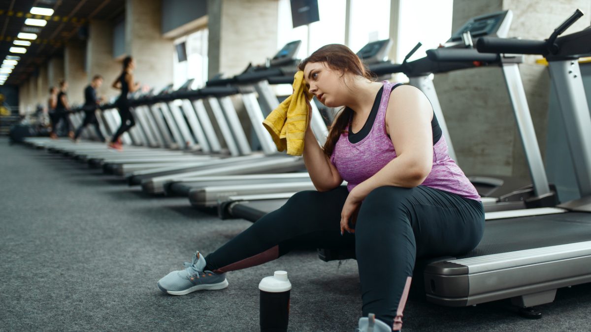 woman sitting down on treadmill after exercise, tired, wiping her head with a yellow towel