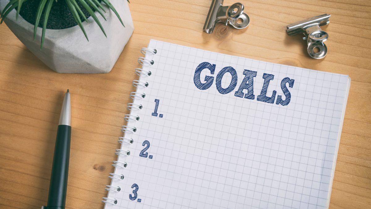 A spiral notebook page titled 'Goals' with a numbered blank list, next to a pen and a small potted plant