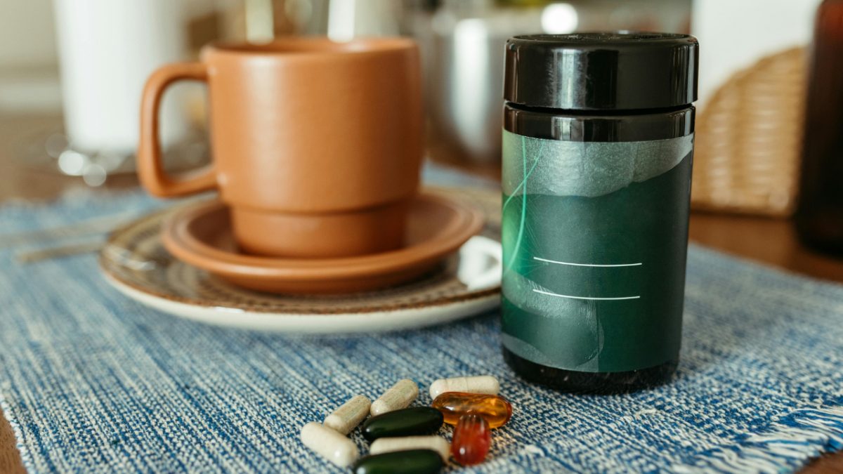 A bottle of supplement capsules next to a scattering of pills and a cup of coffee on a table